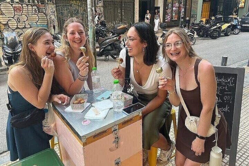 Four people sitting at a table with food and drinks, a chalkboard with writing, and a street scene with scooters and buildings in the background. 1