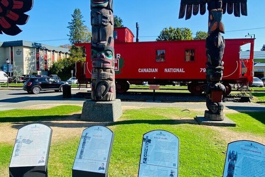A red Canadian National train, totem poles, and informational plaques in a grassy area. 2