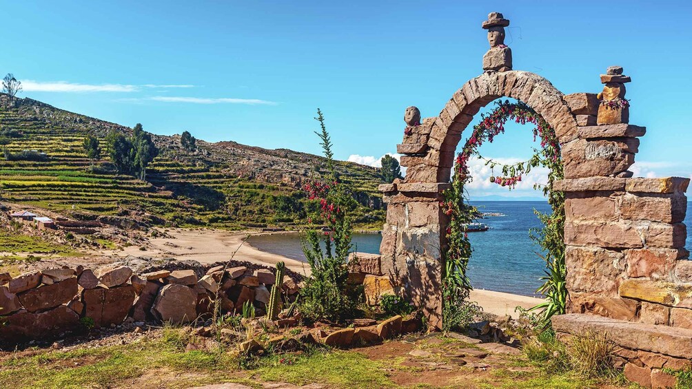 A stone archway with flowers, a beach, terraced fields, and a body of water. 2