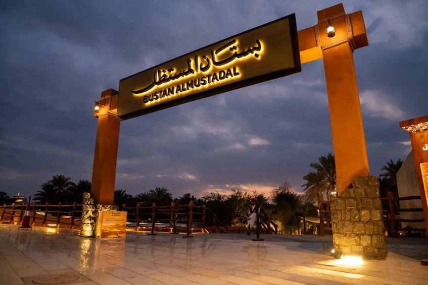 A sign with 'Bustan Almustal' written on it, surrounded by a well-lit area with palm trees and a stone structure. 1