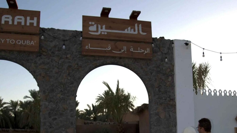 An archway with signs in Arabic and English, palm trees, and a clear sky. 1