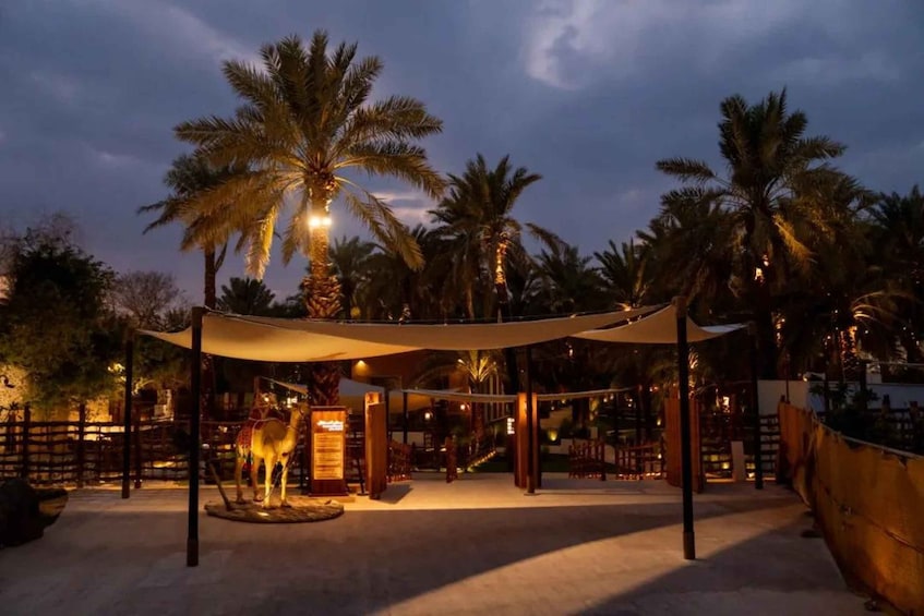A nighttime outdoor area with palm trees, a covered seating area, and a camel statue. 4