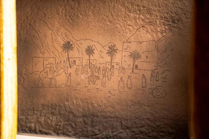 A cave wall with ancient drawings of people, palm trees, and buildings. 5