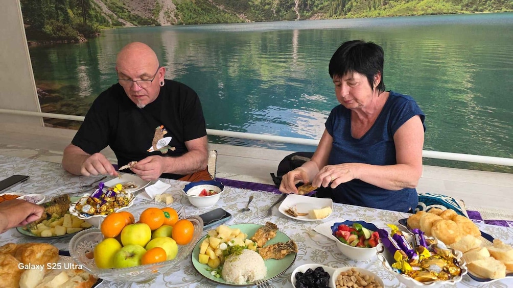 Two people dining at a table with a lake view, plates of food, and a Galaxy S25 Ultra phone. 2