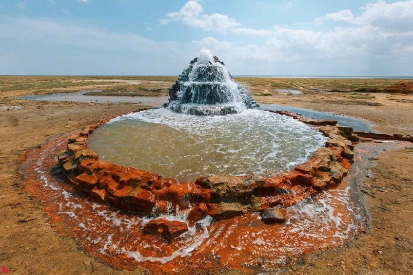 A natural water feature with a central waterfall, surrounded by a red-bedded area, set in a flat, open landscape. 1
