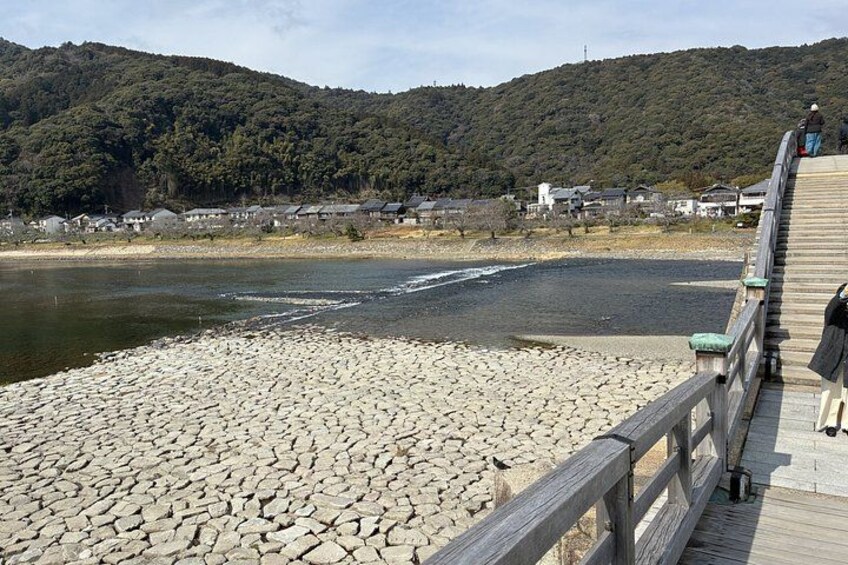 A wooden bridge over a river with a rocky shore, surrounded by hills and houses. 3