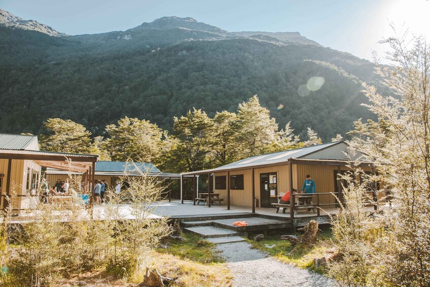 A rustic cabin with a wooden deck and a picnic table, surrounded by a forested mountain backdrop. 2