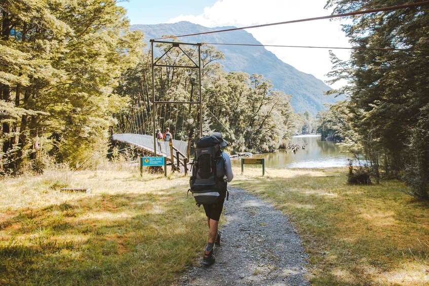 A hiker on a gravel path with a backpack, surrounded by trees and a lake, with mountains in the background. 5