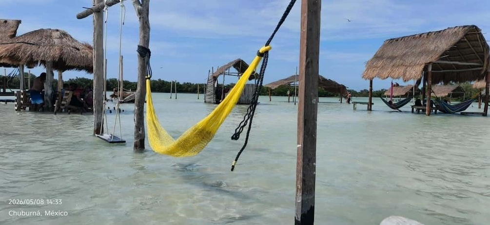 A hammock suspended between wooden posts in a shallow water body, surrounded by thatched-roof structures. 1
