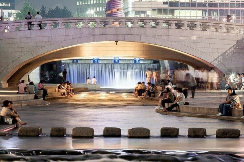 A public square with a stone bridge, a fountain, and people sitting and walking. 5