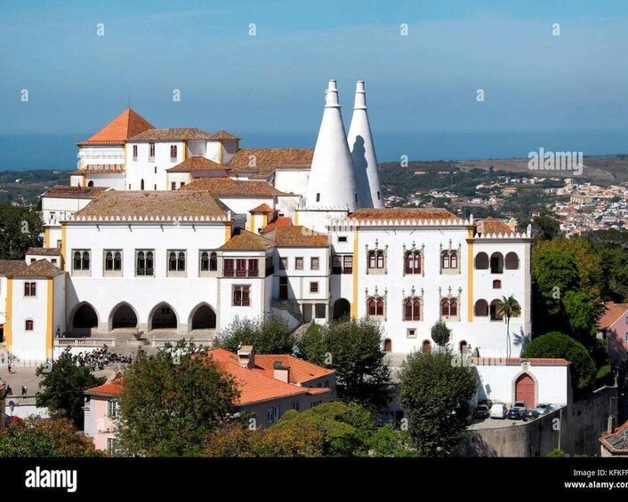 A historic white building with red-tiled roofs and two white conical towers, surrounded by greenery and other buildings. 2