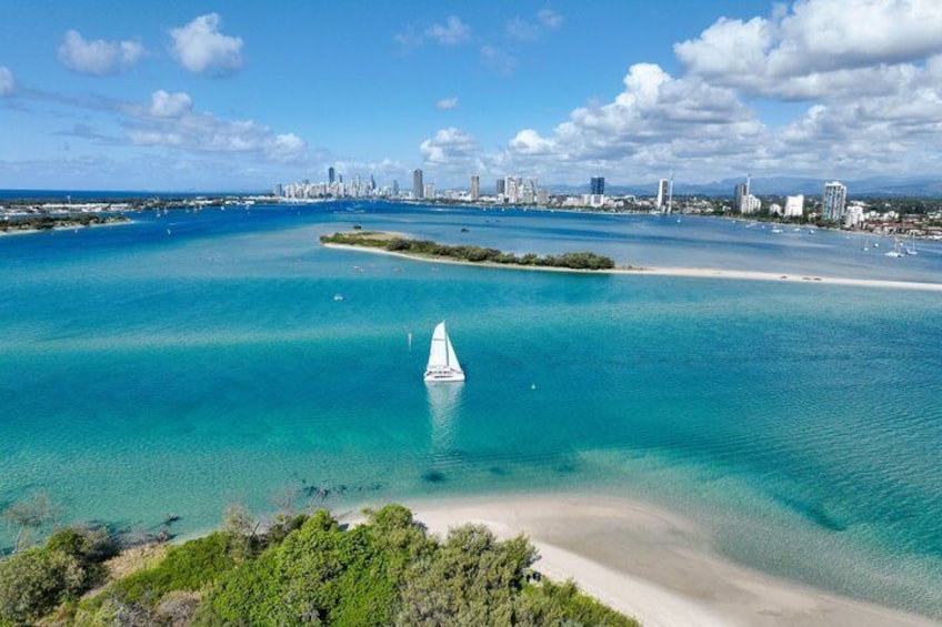 Un paysage urbain côtier avec un voilier sur des eaux turquoise, une plage de sable et, au loin, la silhouette d’une ville. 1