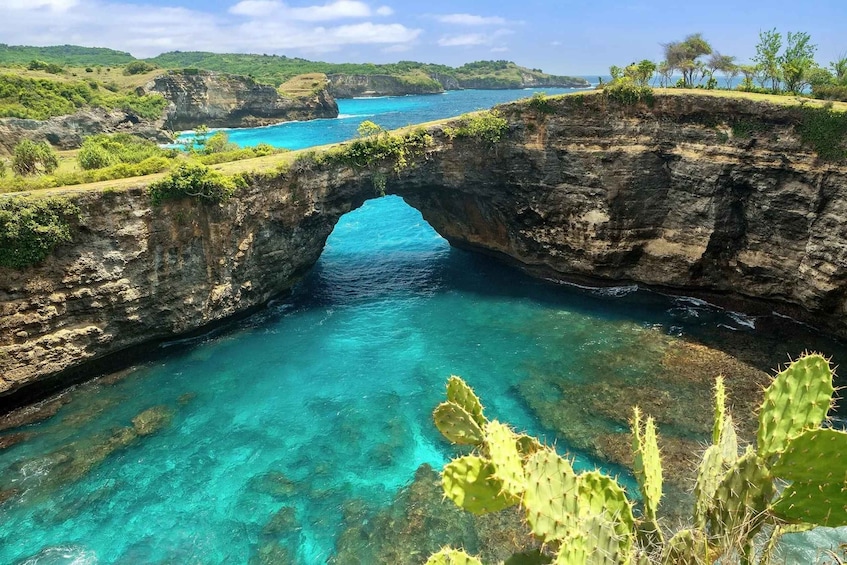 A natural rock arch over a clear blue sea, surrounded by greenery and hills. 2