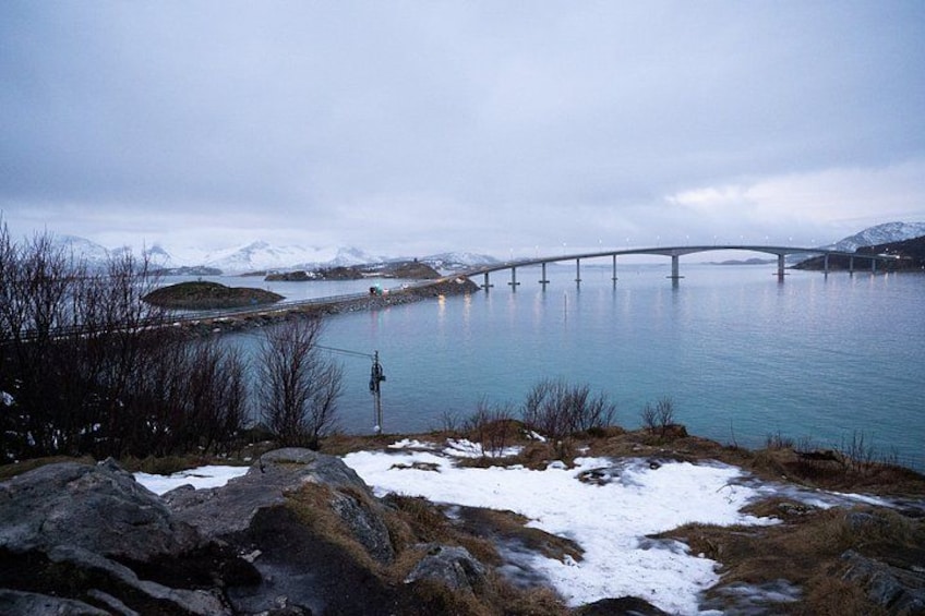 Un ponte sopra acque calme, terreno innevato e una persona in piedi su una roccia. 3