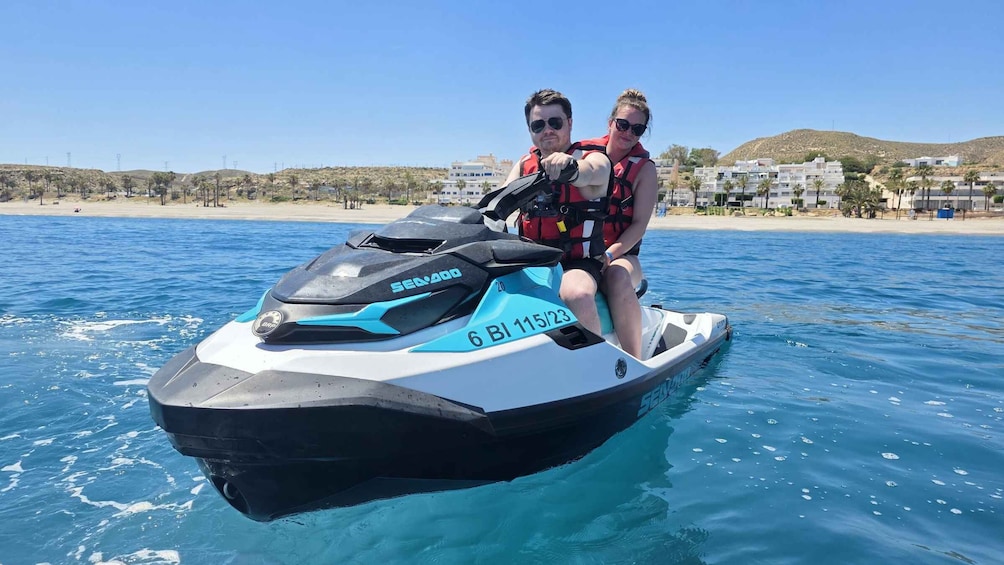 A jet ski with two people on it, a beach, and buildings in the background. 3