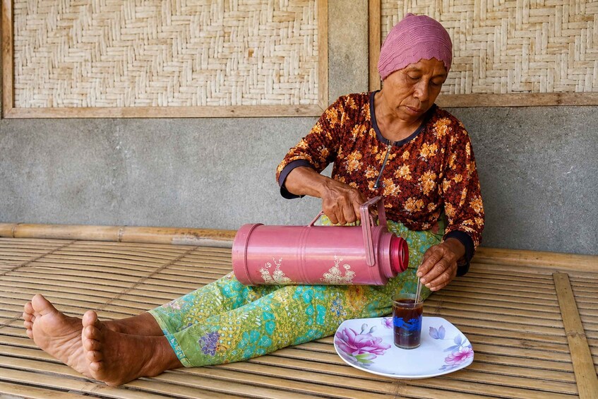 An elderly person sitting on a bamboo mat, pouring a drink from a pink thermos into a glass. 1