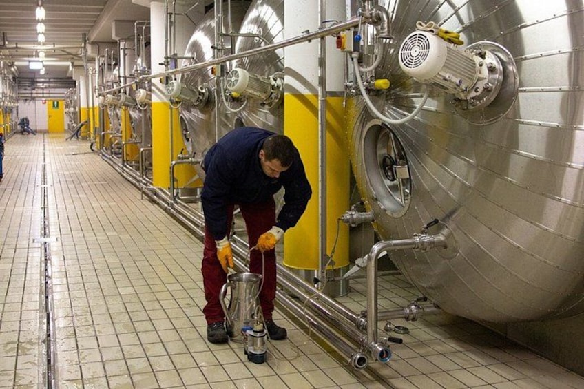 A person in a blue jacket and red pants working with a large stainless steel tank in a facility with yellow walls and a tiled floor. 4