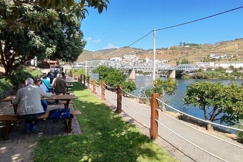 People sitting at picnic tables by a riverside, a bridge in the background, and a hilly landscape. 3
