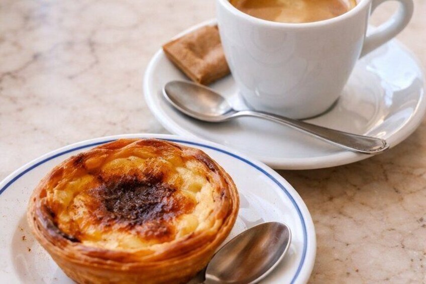 A Portuguese custard tart on a plate with a cup of coffee and a spoon on a table. 4