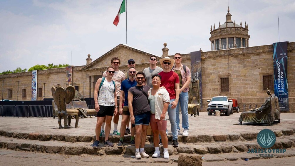 A group of people posing in front of a historic building with a flag, a statue, and banners. 1