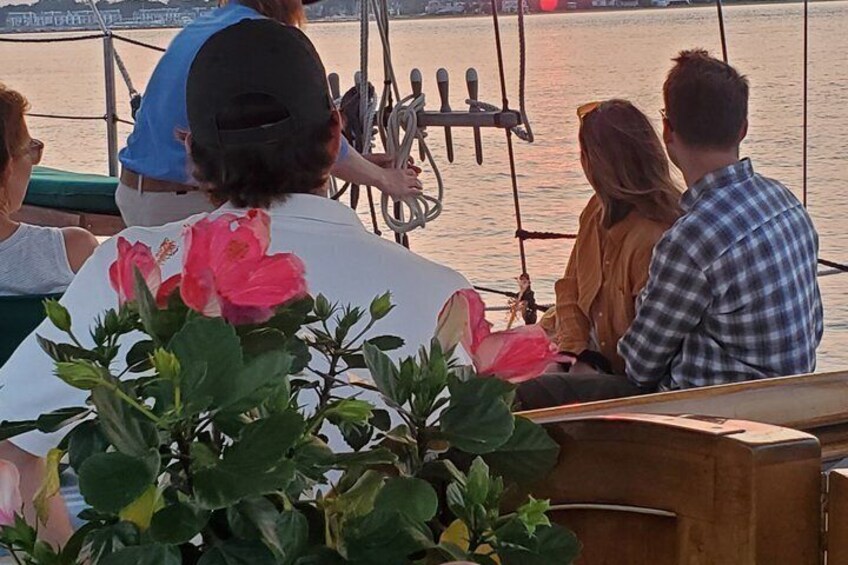 A group of people on a boat with a rope system, a wooden bench, and a plant with pink flowers. 1