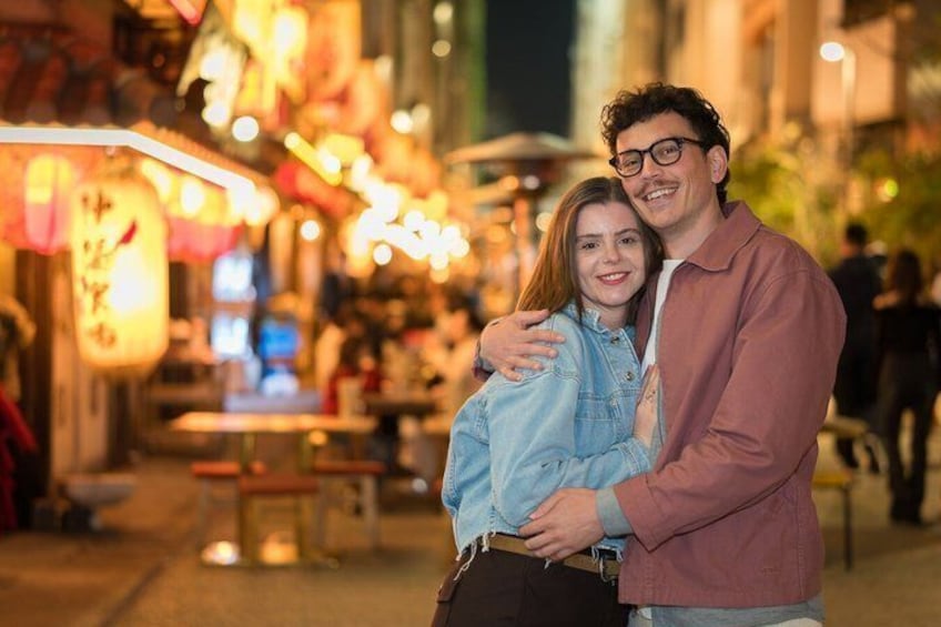 Un couple s’embrasse dans une rue bien éclairée, où les enseignes et les devantures de magasins sont illuminées. 1