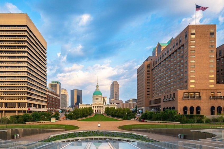 A cityscape with a prominent building with a green dome, surrounded by modern high-rise buildings and a large reflective water feature in the foreground. 2