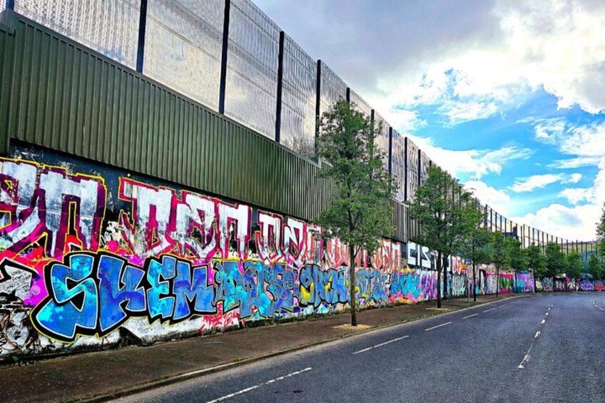 A road with graffiti on a wall, trees, and a clear sky. 1