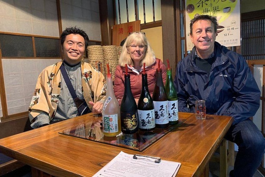 Tres personas sentadas en una mesa de madera con botellas de sake japonés y un menú. 1