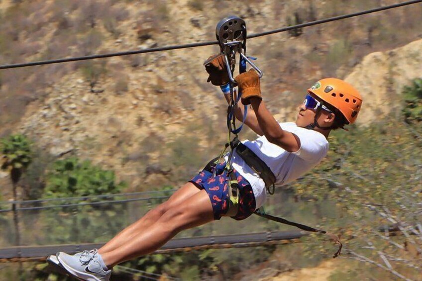 A person zip-lining with a helmet and harness, wearing a white shirt and shorts, against a mountainous backdrop. 2