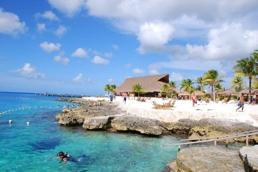 Una playa con agua turquesa cristalina, un rompeolas rocoso, palmeras y un edificio con techo de paja. 2