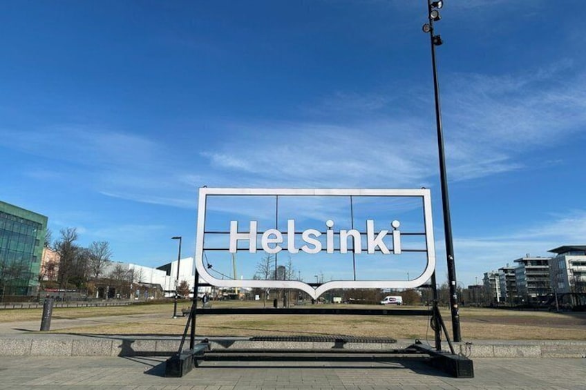 A large sign with 'Helsinki' written on it, set against a clear blue sky with buildings in the background. 5