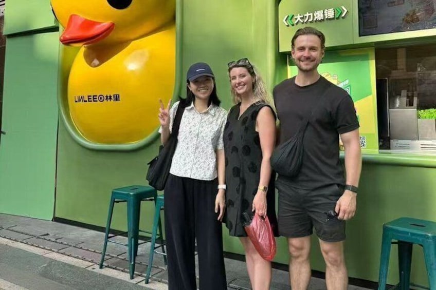 Drei Personen stehen vor einem grünen Gebäude mit einer großen, gelben Enten-Maskottchen und einem Schild mit chinesischen Schriftzeichen. 1