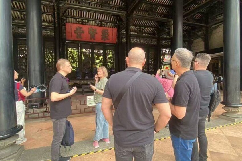 Eine Gruppe von Menschen steht in einem überdachten Pavillon mit traditioneller Architektur, darunter Säulen und ein rotes Banner. 5