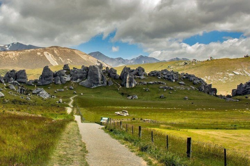 A gravel path leading through a grassy field with rocky outcrops and mountains in the background. 1