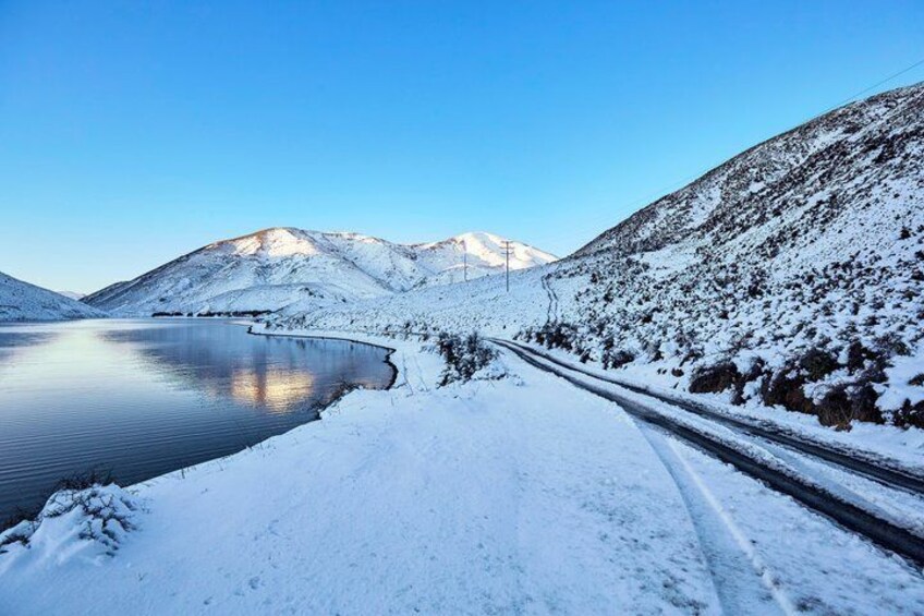 A snow-covered road beside a calm lake with mountains in the background. 5