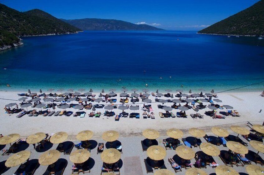 Una spiaggia con file di lettini e ombrelloni, un mare blu cristallino e un paesaggio collinare sullo sfondo. 1