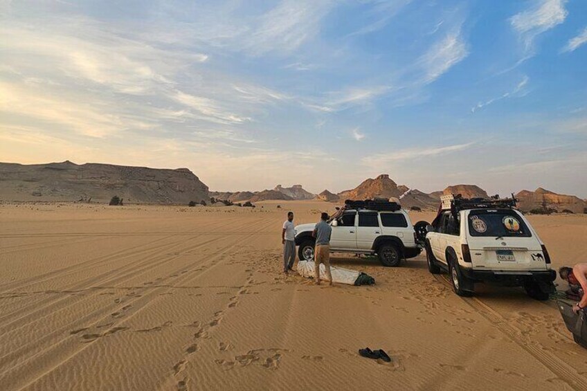 Un paysage désertique avec deux SUV blancs garés sur le sable, une personne debout près de l’un des véhicules, et des empreintes de pas qui s’enfoncent dans le lointain. 2