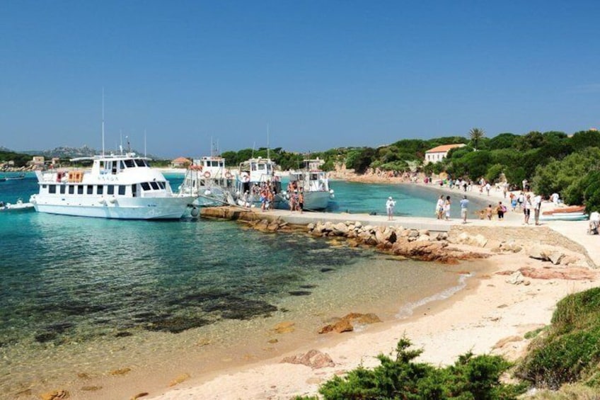 Una zona costera con un barco amarrado en un muelle, agua cristalina, una playa arenosa y gente caminando por la orilla. 3