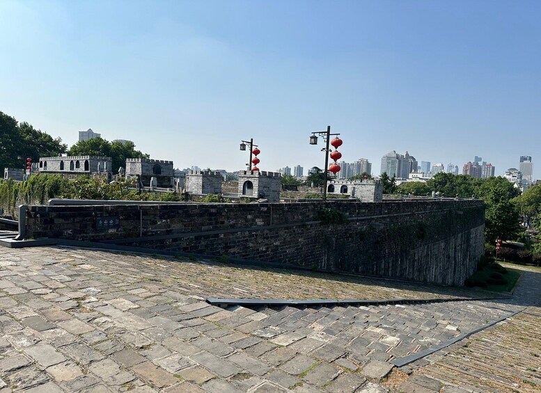 A stone wall with red lanterns, a paved area, and a cityscape in the background. 2