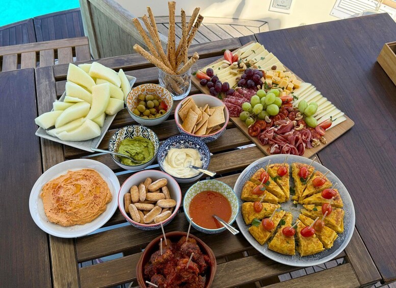 A variety of food items including cheese, olives, dips, and a pizza on a wooden table by a pool. 2