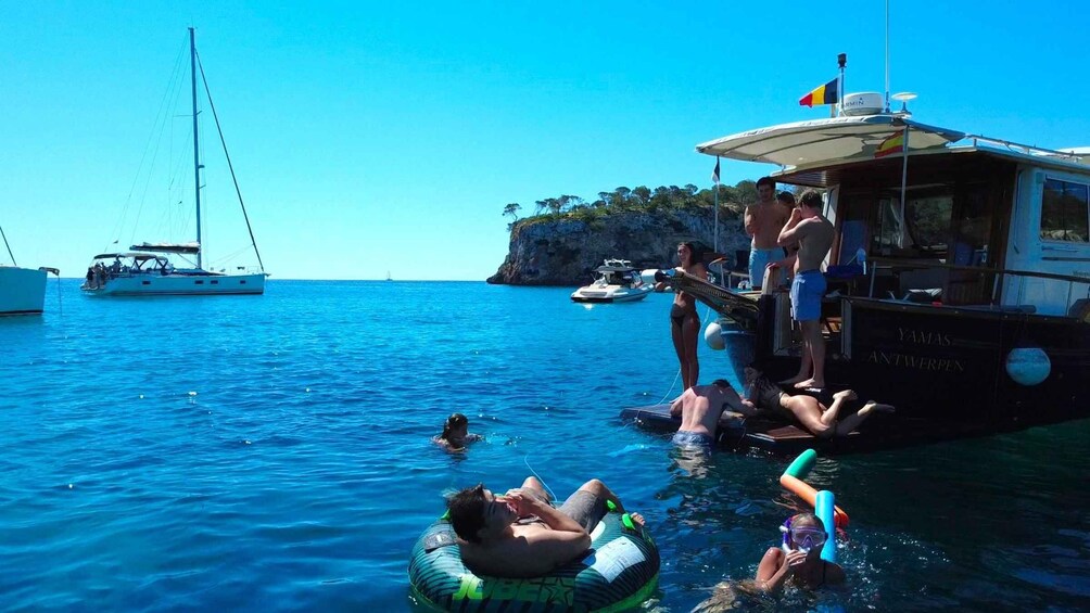 A group of people swimming and floating on a boat in a clear blue sea, with another sailboat in the background. 4
