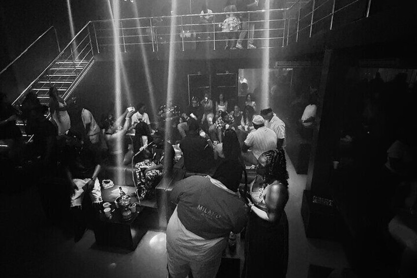 A crowded indoor event with people standing and sitting, bright lights, and a staircase. 3