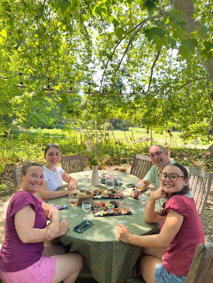 A group of people sitting at an outdoor table with food and drinks, surrounded by greenery and trees. 3