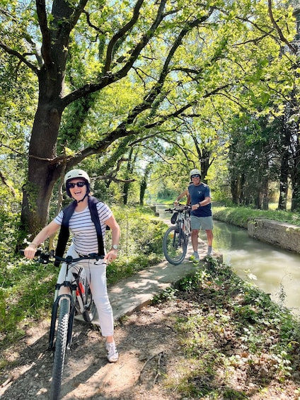 Two people riding bicycles on a tree-lined path beside a canal. 1
