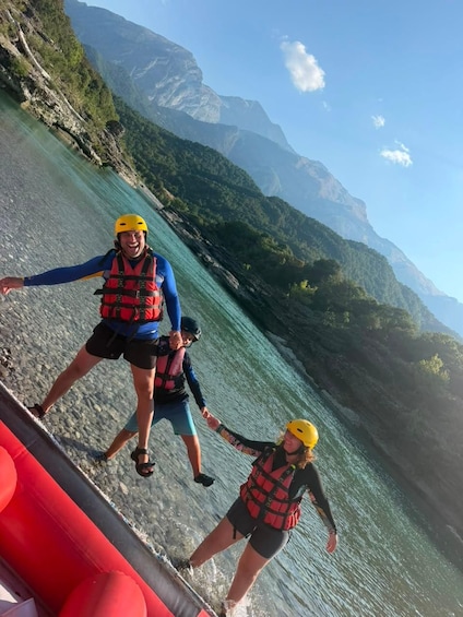 Two people in life jackets and helmets walking on a rocky shore by a body of water with mountains in the background. 3