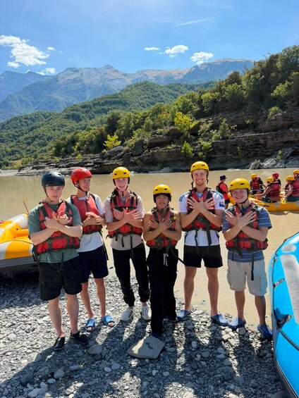 A group of people wearing helmets and life vests standing on a rocky shore with a blue inflatable raft nearby. 5