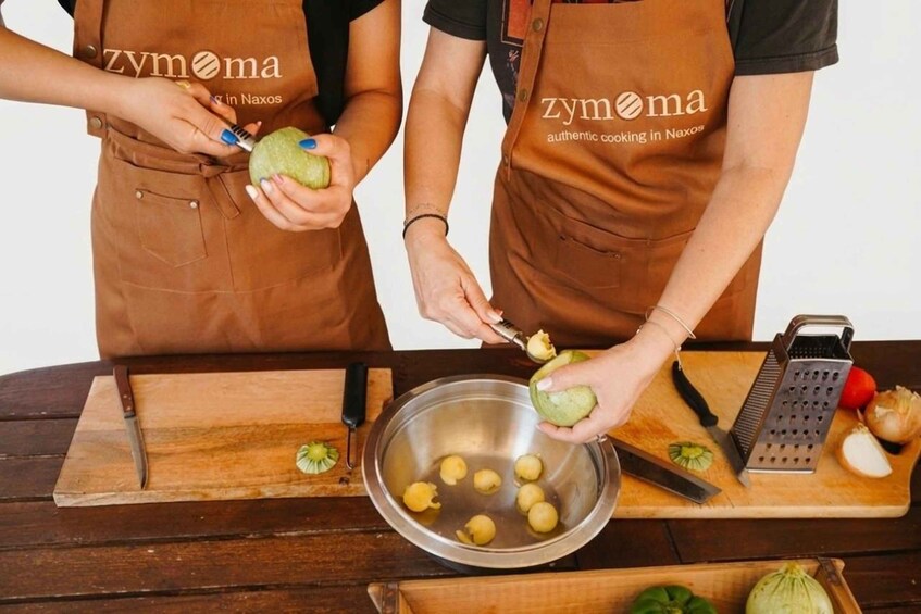 Two people in aprons preparing food, with a bowl of peeled potatoes and a grater on the table. 3