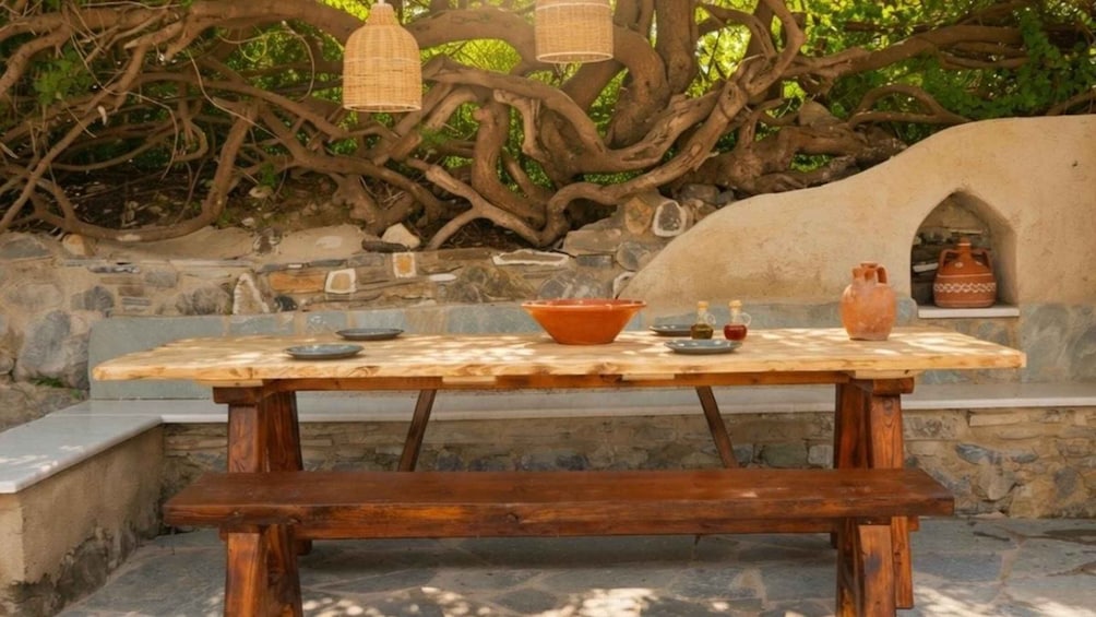A rustic outdoor dining table with a bowl, a jug, and a candle, set against a stone wall and tree roots. 1