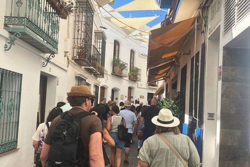 A narrow street with people walking, white buildings with balconies, and a blue and yellow awning. 3